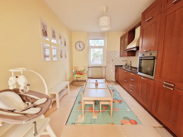 Helle Küche mit Holzschränken, Kindertisch und Stühlen auf einem bunten Teppich.
