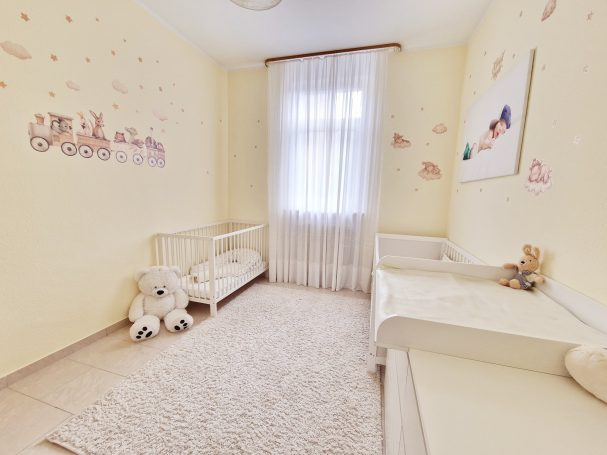 Helles Kinderzimmer mit beigen Wänden, Teppich, einem Fenster und Spielzeug.