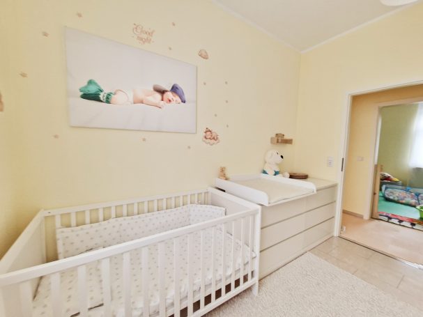 Helle Kinderzimmer mit Gitterbett, Wickelkommode und einem Gemälde an der Wand.